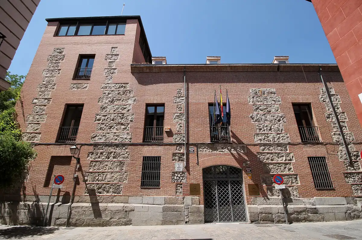 Fachada de un palacio urbano de piedra y ladrillo de tres alturas, con portada adintelada, rejería en los balcones y un escudo sobre la puerta principal, en una calle estrecha del centro de Madrid.