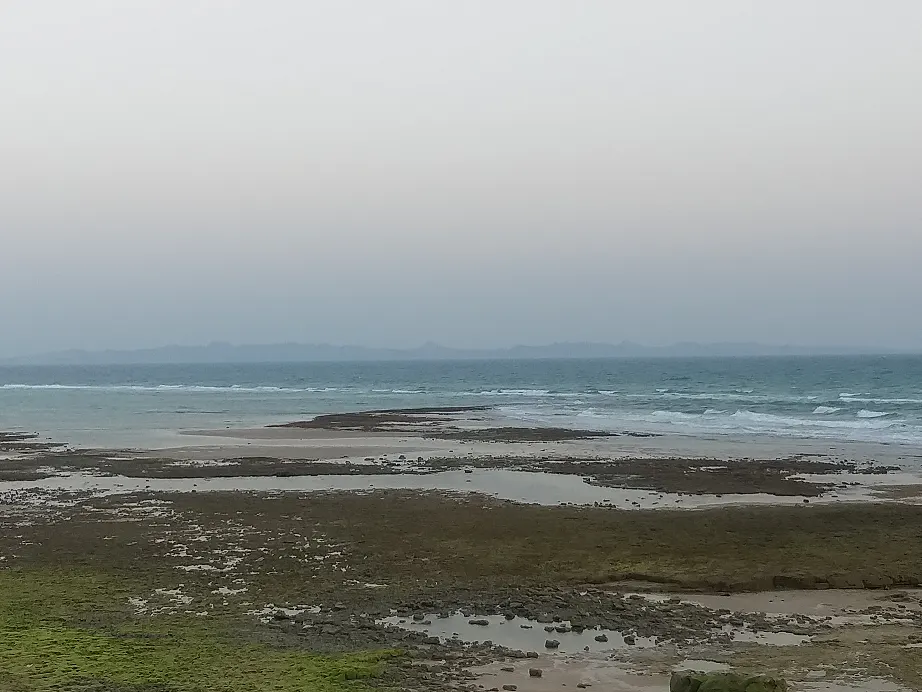 Costa de arena y roca al descubierto con la marea baja; tras una franja de mar turquesa se recorta en el horizonte, entre la calima, la silueta montañosa de una isla.