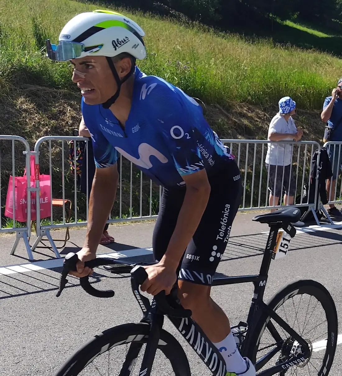 Enric Mas, con casco blanco y maillot azul del Movistar Team, pedalea sobre una bicicleta negra por una carretera flanqueada por vallas y espectadores, con un talud de hierba al fondo.