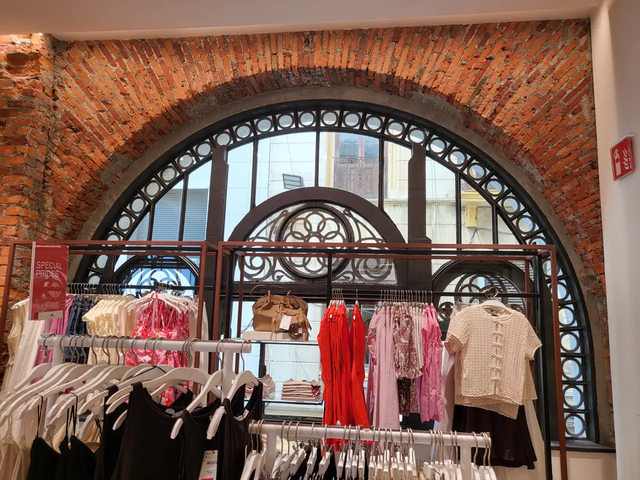 Interior de una tienda de ropa con percheros de blusas y vestidos en blanco, rojo y rosa ante un gran ventanal de arco con celosía de hierro forjado y muro de ladrillo visto; un cartel anuncia «special prices».