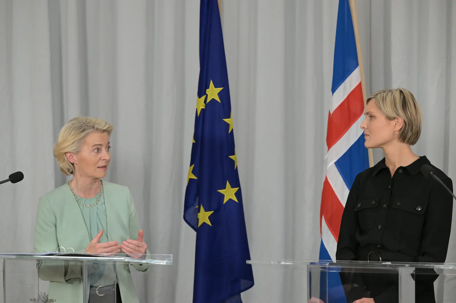 Dos mujeres hablan en atriles de metacrilato ante una cortina gris; entre ellas, la bandera de la Unión Europea y la de Islandia. La de la izquierda gesticula mientras habla; la de la derecha la escucha girada hacia ella.