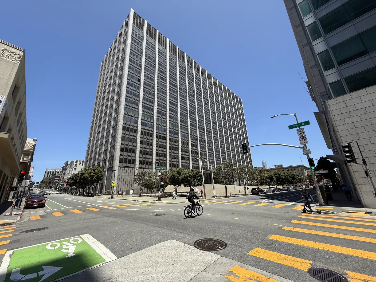 Torre federal de veinte plantas con fachada de ventanas oscuras y nervios verticales de piedra clara, vista desde el cruce de dos calles con pasos de peatones amarillos, un ciclista y un patinete en primer plano, bajo cielo despejado.