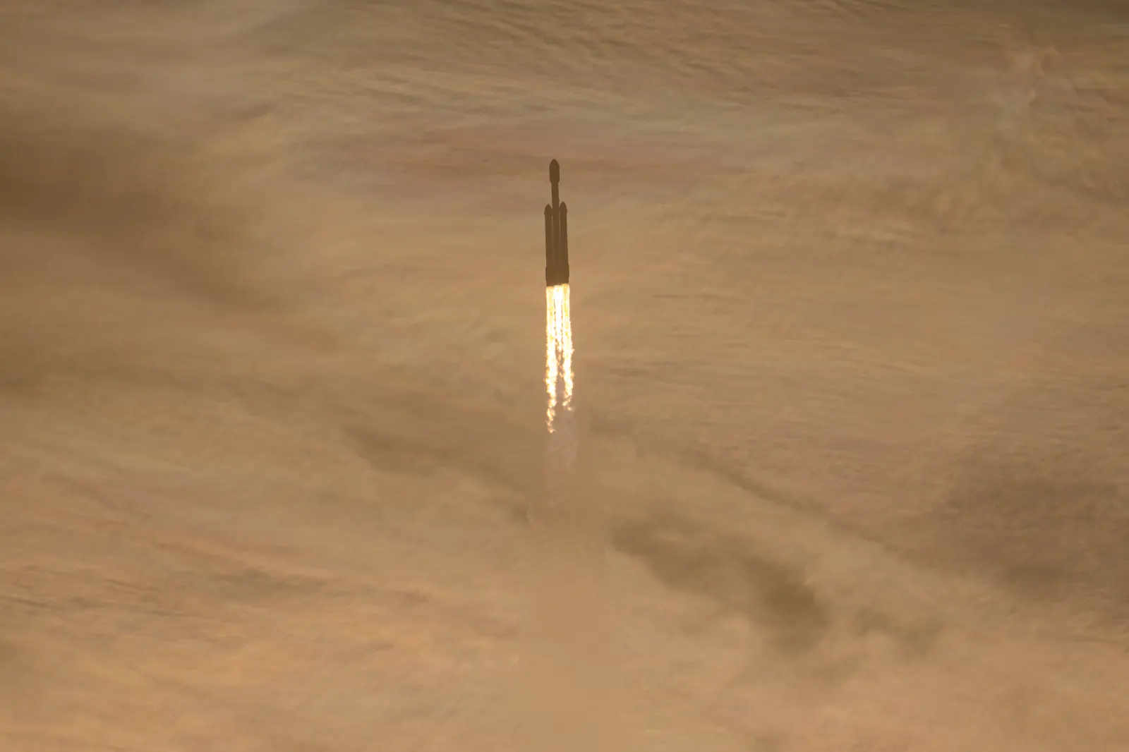 Silueta oscura de un cohete Falcon Heavy ascendiendo entre nubes teñidas de naranja, con dos columnas de fuego blanco bajo los propulsores