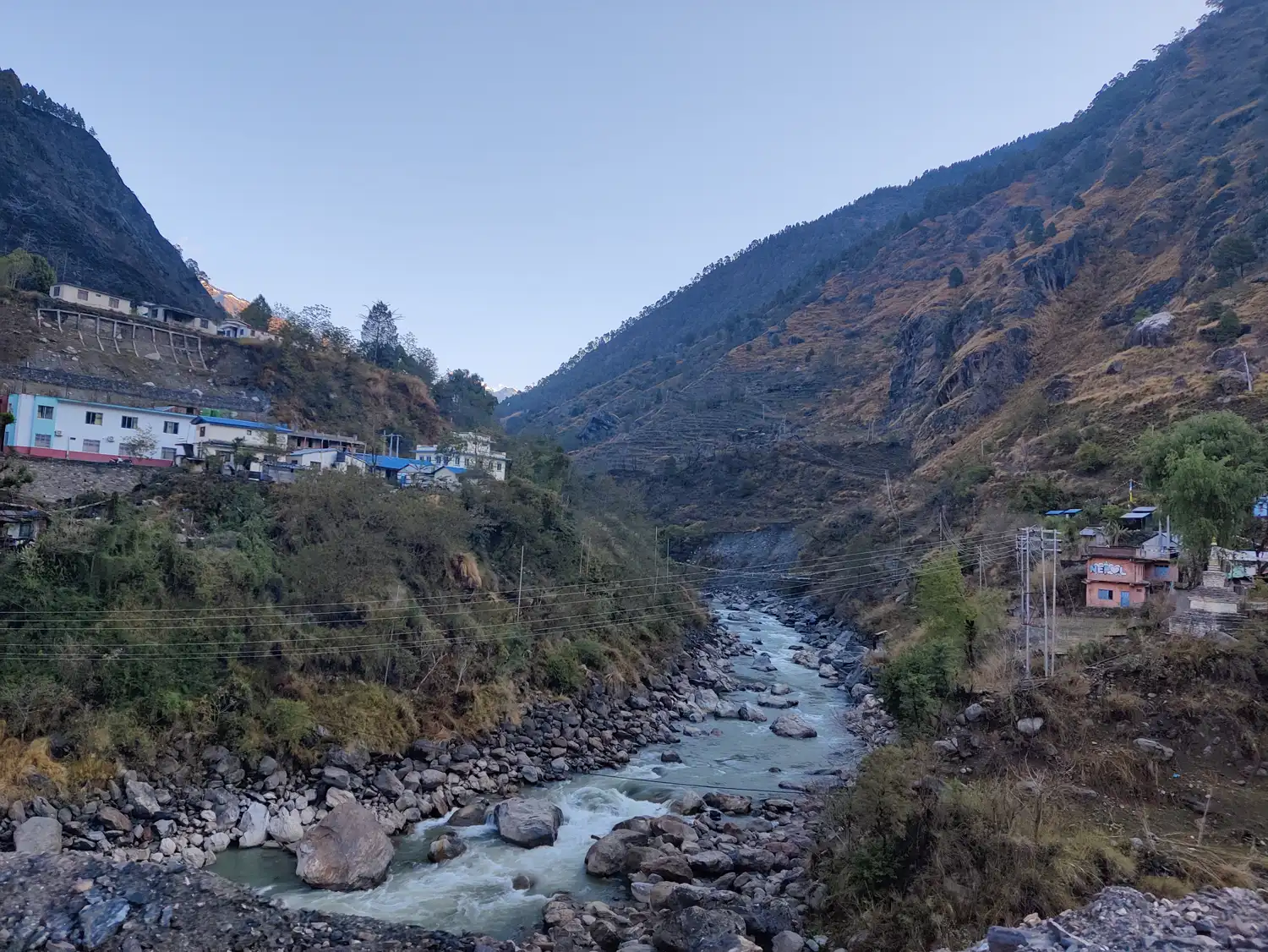 Valle estrecho de montaña con un río de aguas claras entre grandes rocas; en las dos laderas, casas de varias plantas, tendido eléctrico y vegetación seca