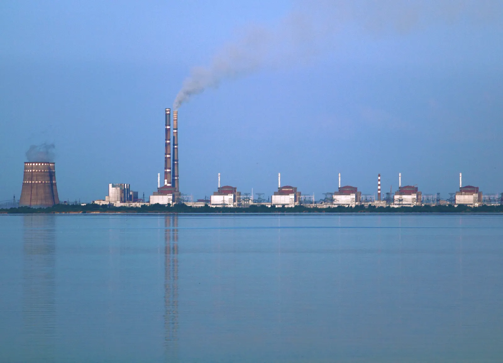 Vista de la central nuclear de Zaporiyia desde la otra orilla del embalse de Kajovka: la fila de seis edificios de reactor a la derecha y, a la izquierda, la torre de refrigeración y la chimenea de la central térmica vecina, reflejadas en el agua.