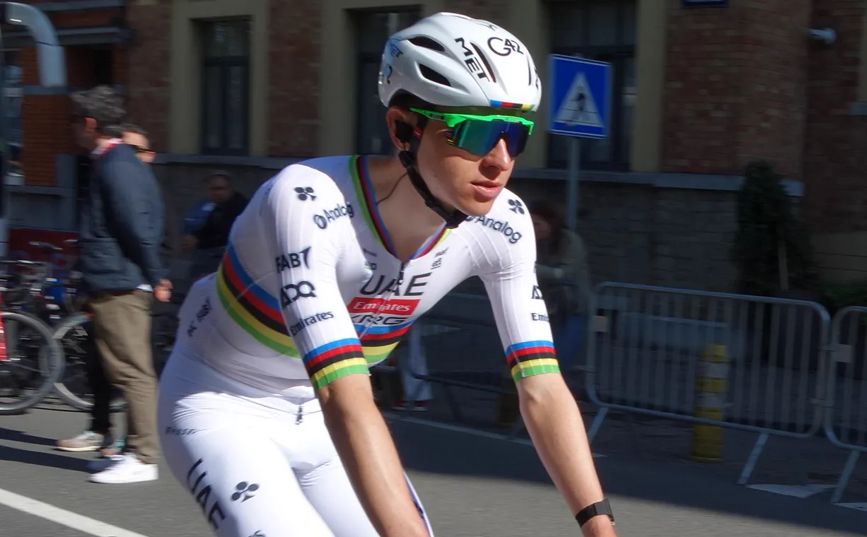 Tadej Pogacar, con casco blanco, gafas verdes y maillot arcoíris de campeón del mundo del UAE Team Emirates, rueda sobre el asfalto junto a unas vallas con público detrás.