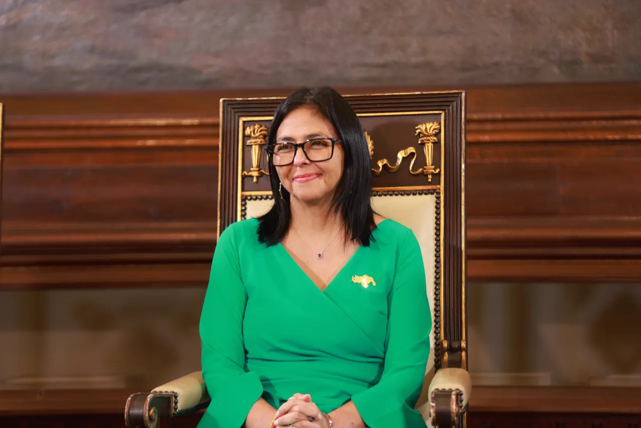 Delcy Rodríguez, sentada en un sillón dorado con vestido verde y un prendedor con el mapa de Venezuela, en un salón de madera