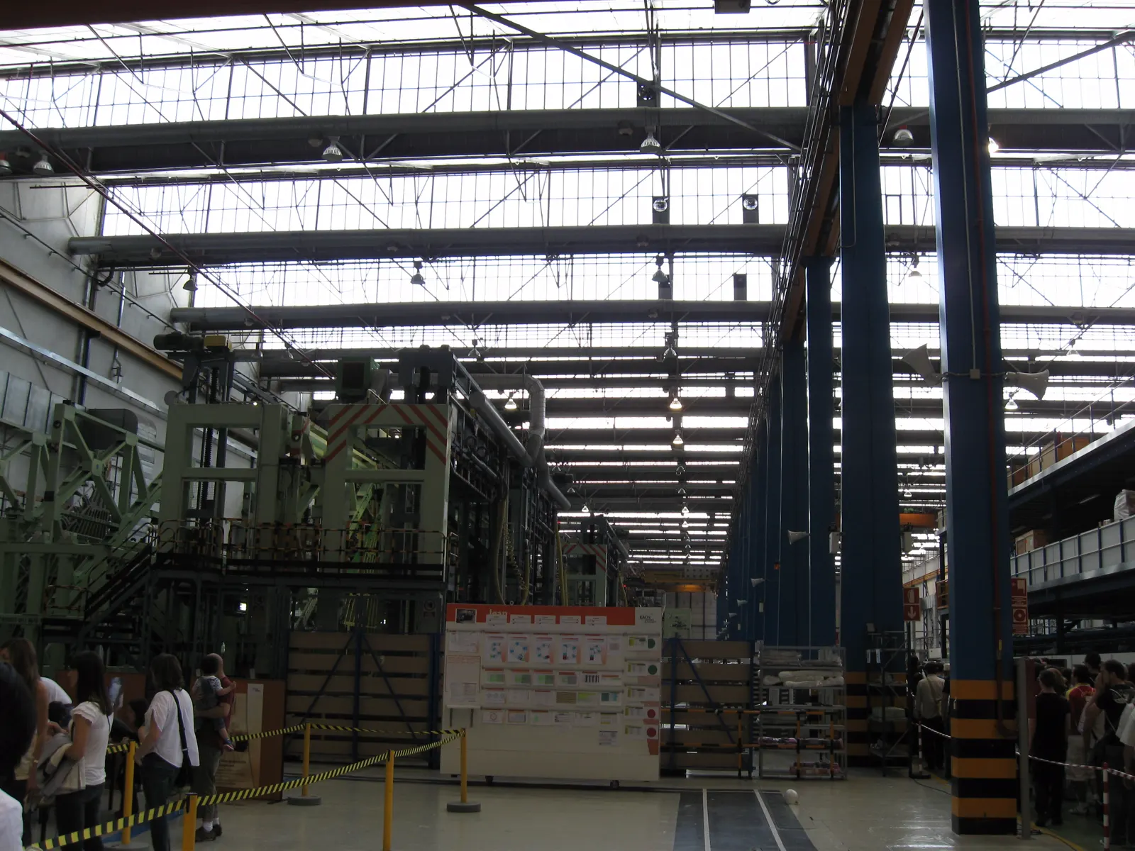 Interior de una nave industrial de gran altura con estructura metálica y lucernarios en el techo, maquinaria de montaje verde a la izquierda, pilares azules a la derecha y un grupo de visitantes tras un cordón de seguridad en primer plano.
