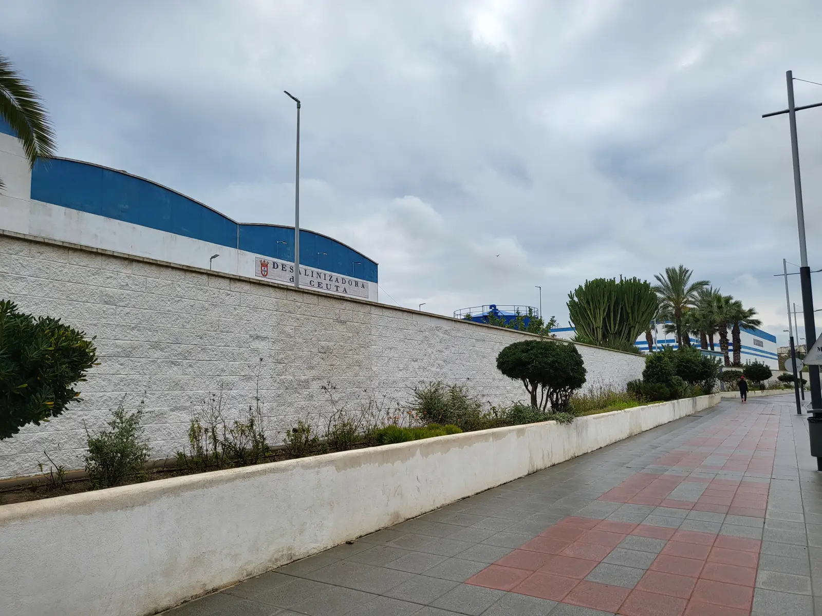 Muro blanco de sillería junto a un paseo peatonal con losetas rojas y grises; tras el muro asoman las naves industriales de cubierta azul de la planta desalinizadora y un cartel de piedra con el escudo de la ciudad que reza «Desalinizadora de Ceuta». Cielo nublado y palmeras al fondo.