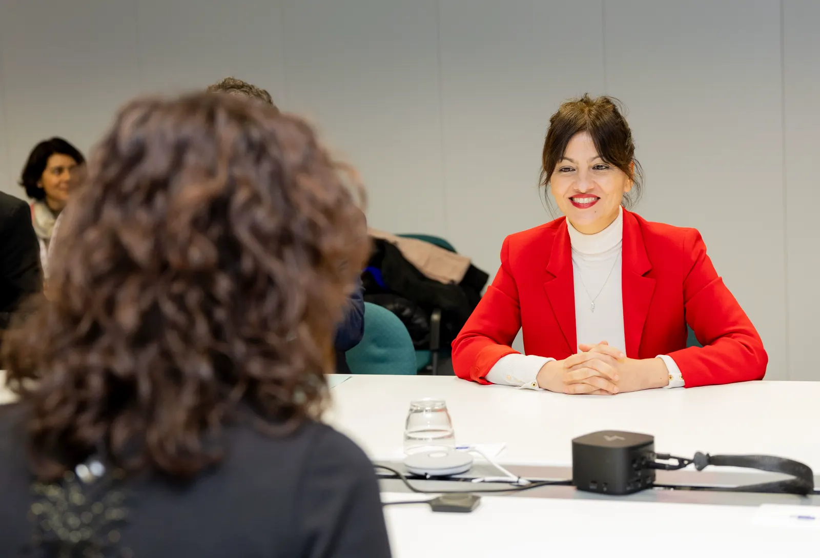 La ministra Sira Rego, con chaqueta roja y jersey blanco de cuello alto, sentada con las manos entrelazadas ante una mesa blanca de reunión y mirando a una interlocutora que aparece de espaldas y desenfocada en primer plano.