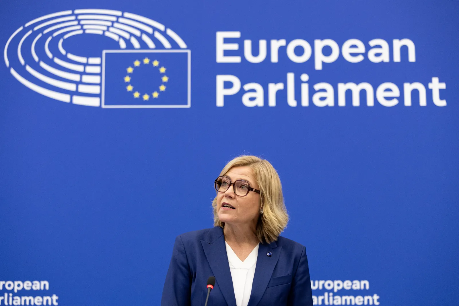 Henna Virkkunen, con gafas y chaqueta azul marino, habla ante un micrófono delante de un panel azul con el logotipo del Parlamento Europeo