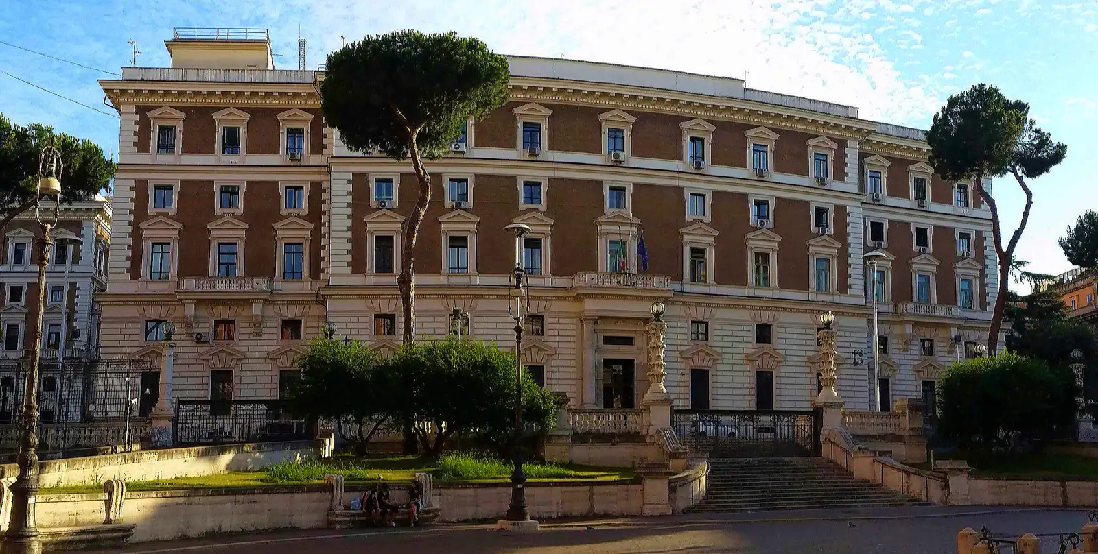Fachada del Palazzo del Viminale, sede del Ministerio del Interior italiano en Roma: un edificio de cuatro plantas con los pisos altos de ladrillo rojo y el zócalo de piedra clara, pinos romanos y farolas de hierro delante, una doble escalinata que sube hasta la puerta principal y, sobre ella, las banderas de Italia y de la Unión Europea en el balcón central.