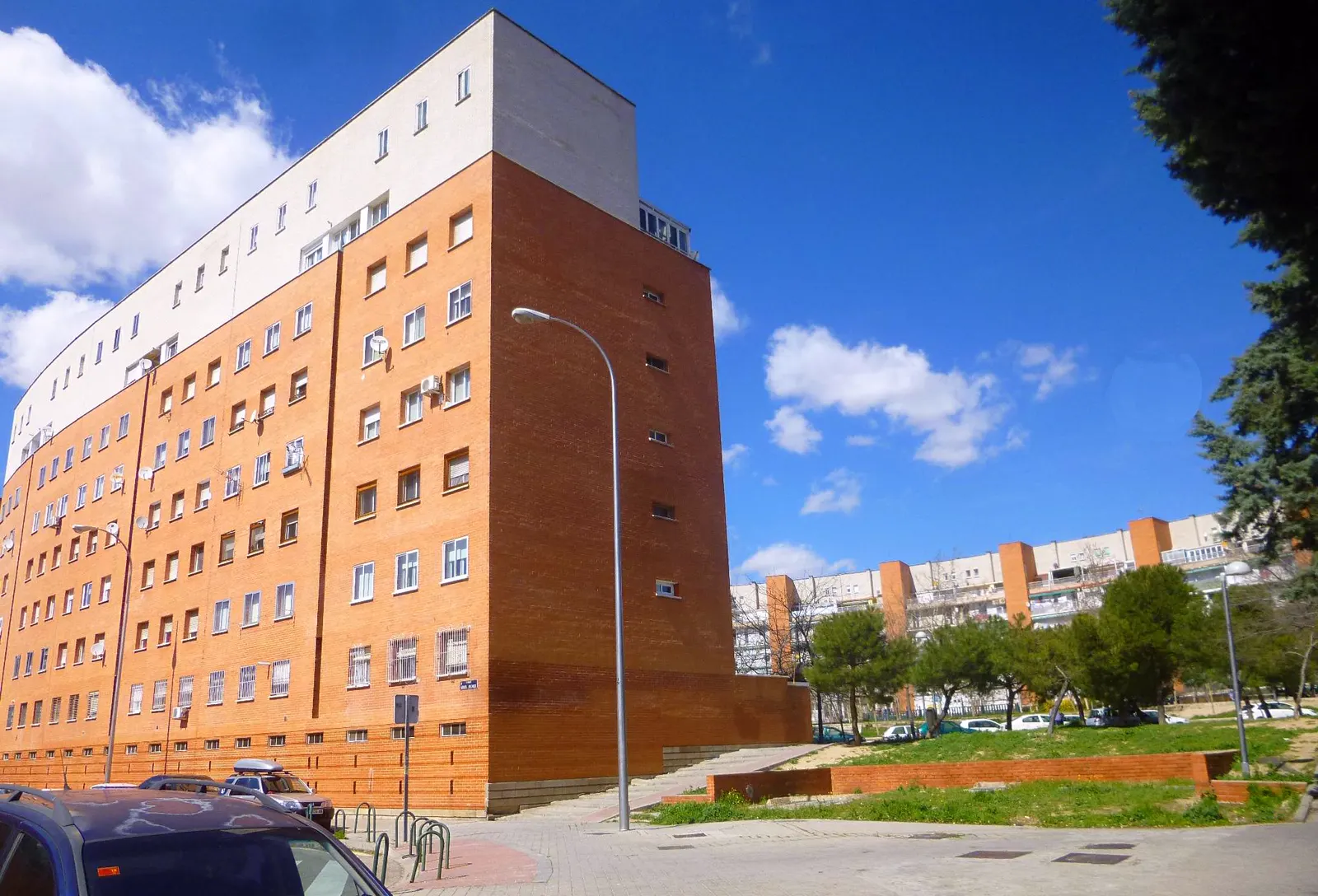 Bloque de viviendas de ladrillo rojo de ocho alturas en el barrio de Moratalaz, en Madrid, con coches aparcados delante y cielo azul con nubes.