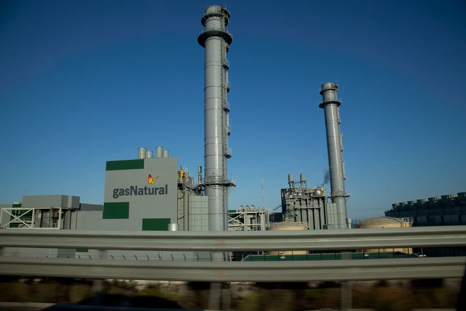 Central eléctrica de ciclo combinado con dos altas chimeneas metálicas contra un cielo azul; en la fachada del edificio principal se lee el rótulo gasNatural y en primer plano cruza la quitamiedos de una carretera