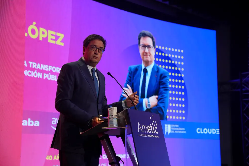 Óscar López, con traje oscuro y gafas, habla ante un micrófono en un atril con el logotipo de Ametic; detrás, una pantalla iluminada en rosa y azul con su apellido y su retrato