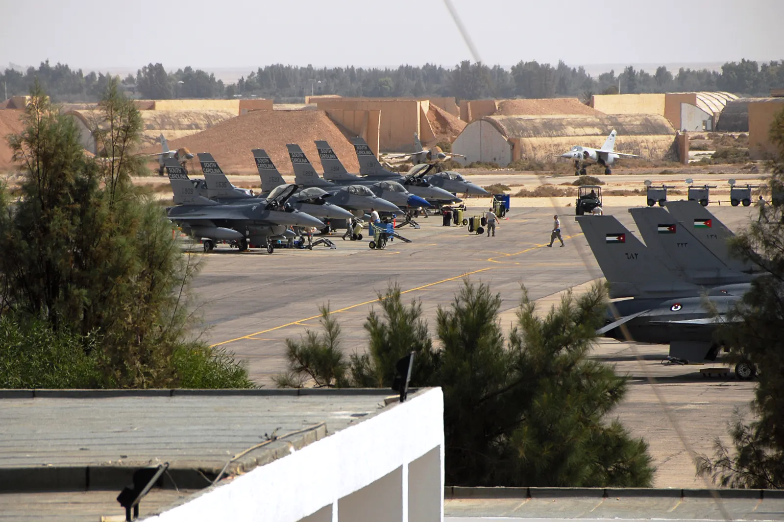 Plataforma de una base aérea en pleno desierto con una decena de cazas F-16 aparcados en fila; a la derecha, tres aviones con la bandera de Jordania pintada en la cola; al fondo, hangares de hormigón semienterrados y un caza rodando por la pista.