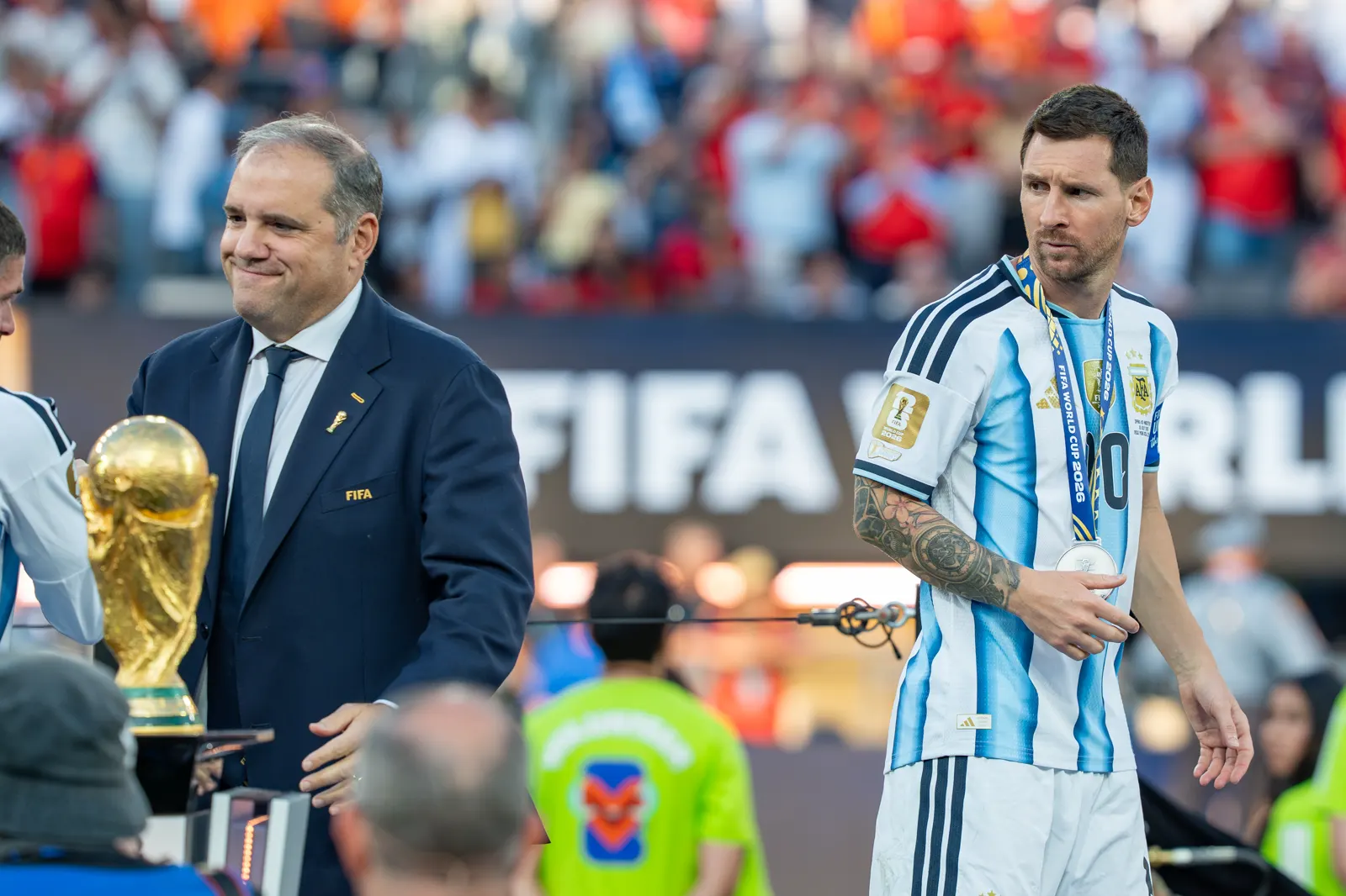 Lionel Messi, con la camiseta albiceleste y la medalla de subcampeón colgada al cuello, mira hacia un lado en el podio; a su izquierda, un dirigente con el escudo de la FIFA en la solapa sonríe junto al trofeo dorado de la Copa del Mundo