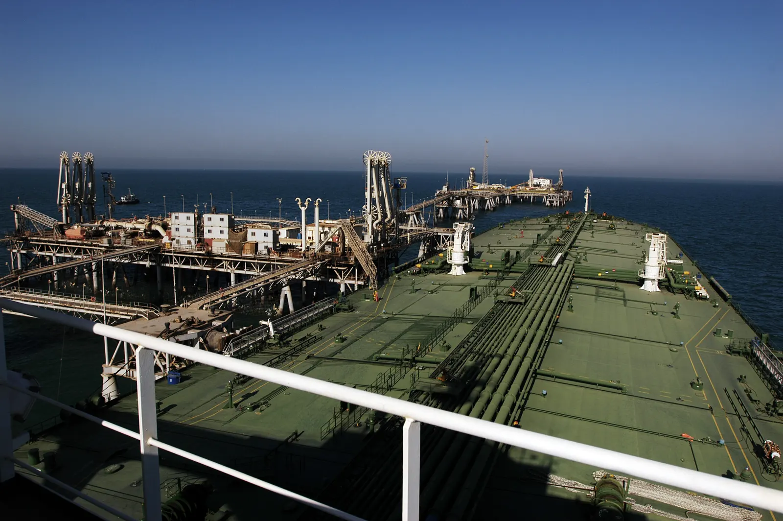 Vista desde el puente de un petrolero: la cubierta verde, con sus tuberías y válvulas, se extiende hacia una plataforma metálica de carga rodeada de mar en calma bajo un cielo despejado.