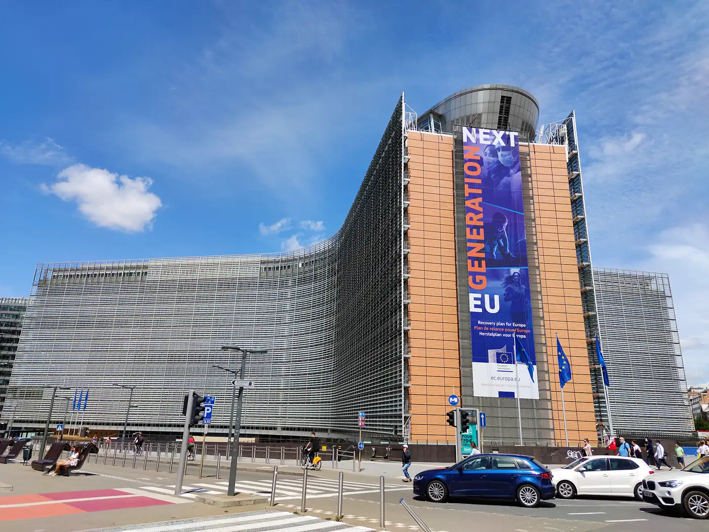 Fachada curva del edificio Berlaymont, sede de la Comisión Europea en Bruselas, con una lona vertical de varios pisos con el lema «NextGenerationEU» y el texto «Recovery plan for Europe»; banderas de la Unión Europea y tráfico en la calle.