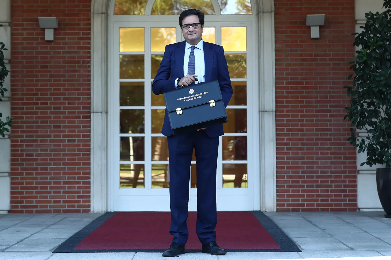 Óscar López, con traje azul marino y gafas, posa de pie sobre una alfombra roja ante la puerta acristalada del palacio de la Moncloa sosteniendo la cartera ministerial rotulada «Ministro para la Transformación Digital y de la Función Pública»