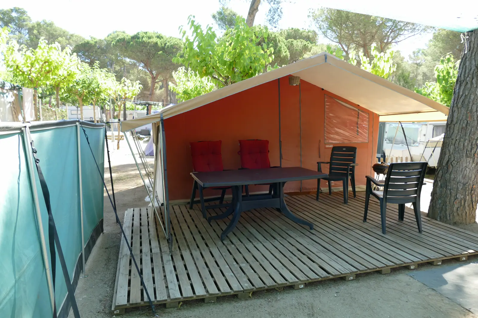 Tienda de campaña grande de lona naranja con porche, mesa y sillas de plástico sobre una tarima de madera en una parcela arbolada de un camping; al fondo, caravanas entre pinos