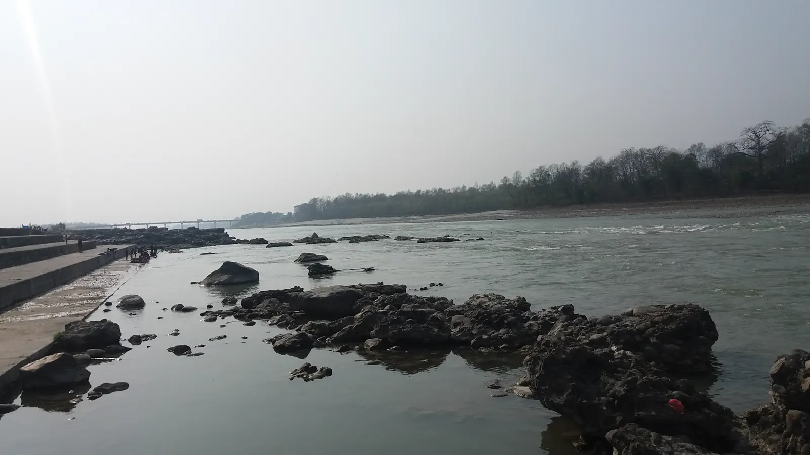 Río ancho de aguas verdosas entre bancos de roca oscura; a la izquierda, un dique de hormigón con figuras diminutas; al fondo, un puente y una orilla boscosa bajo un cielo blanquecino