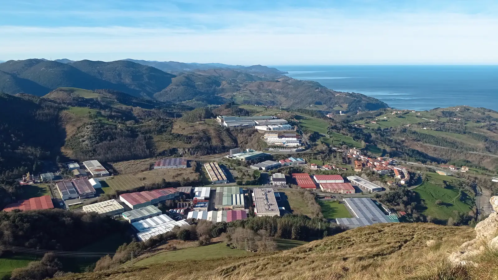 Vista aérea de un polígono industrial en un valle verde: dos decenas de naves rectangulares con cubiertas rojas, grises y verdes rodeadas de prados y bosque, con montes y el mar Cantábrico al fondo.