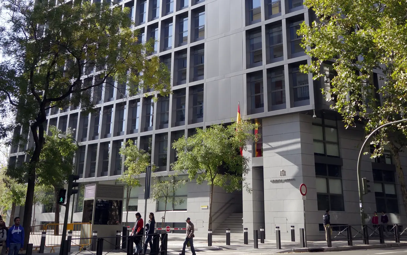 Fachada del edificio de la Audiencia Nacional en Madrid, un bloque moderno de vidrio y paneles grises; junto a la esquina, la bandera de España, el escudo y el rótulo «Audiencia Nacional», con árboles y viandantes en la acera.