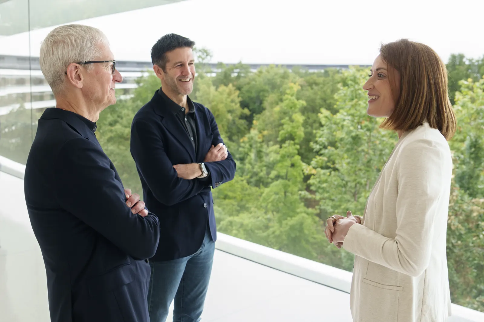 Tim Cook, con gafas y pelo cano, y John Ternus, de brazos cruzados, conversan de pie junto a una barandilla de cristal frente a Roberta Metsola, con arbolado al fondo