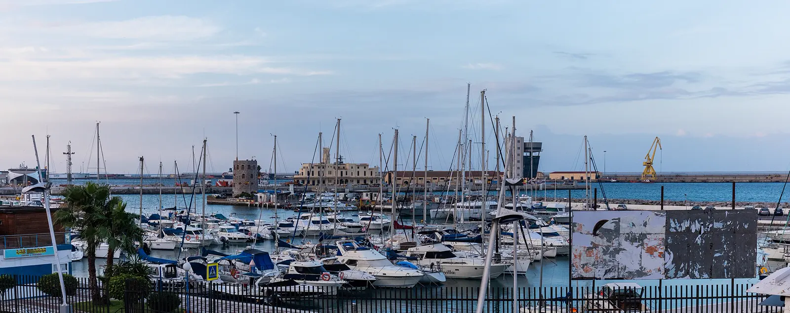 Vista panorámica del puerto de Ceuta al atardecer, con decenas de veleros y embarcaciones deportivas amarradas en primer plano, una grúa amarilla y el dique de abrigo al fondo sobre el mar.