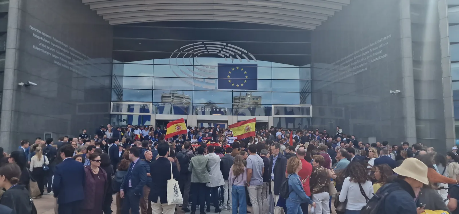 Concentración ante la entrada acristalada del edificio Altiero Spinelli del Parlamento Europeo, con la bandera de la Unión Europea sobre la fachada. Varios centenares de personas, muchas trajeadas y con cordones de acreditación, se agrupan ante la puerta; dos banderas españolas ondean sobre la multitud junto a varias pancartas.