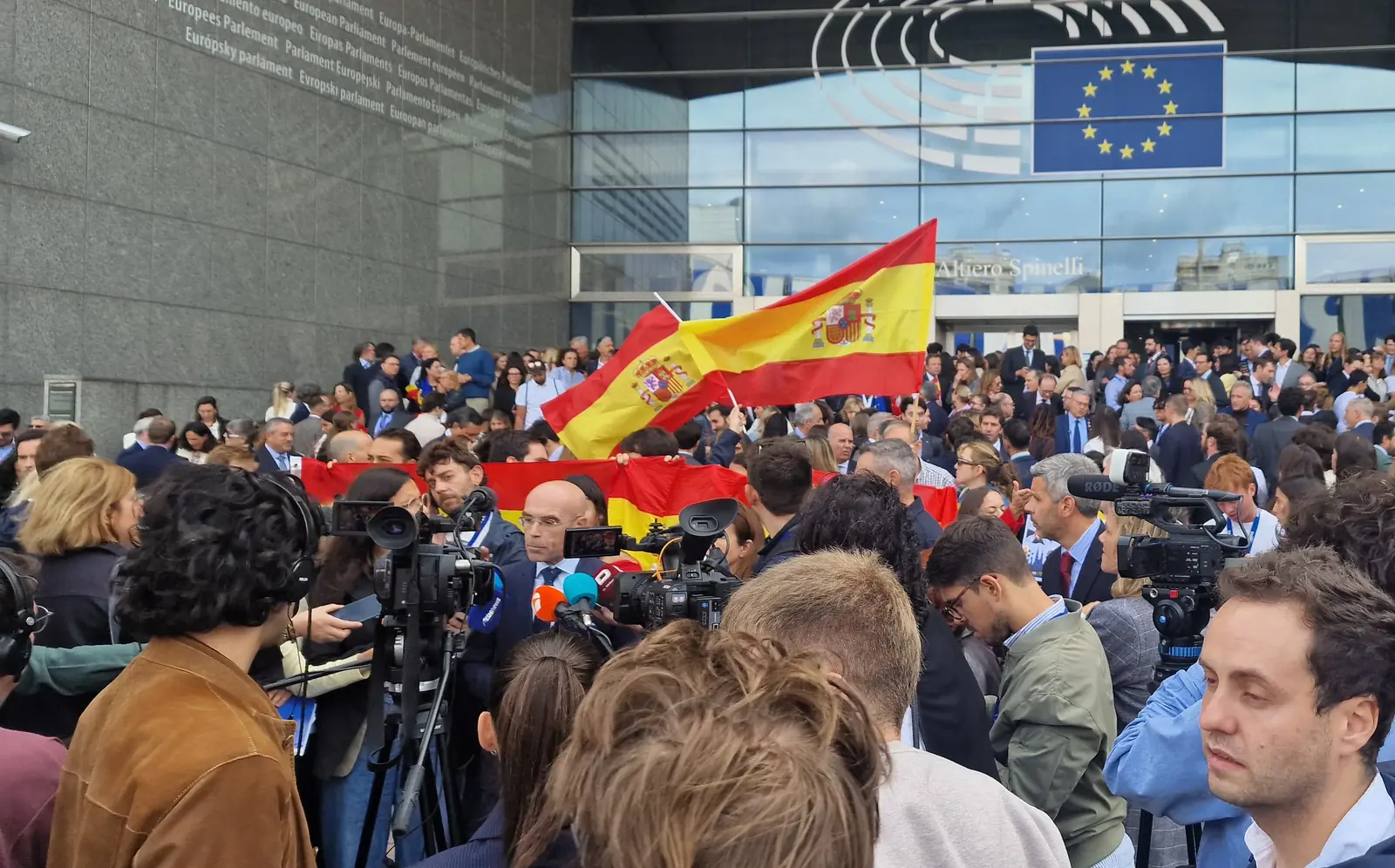Jorge Buxadé, de traje oscuro y corbata azul, responde a media docena de micrófonos ante varias cámaras de televisión, con una bandera de España ondeando detrás y la entrada del edificio Altiero Spinelli al fondo