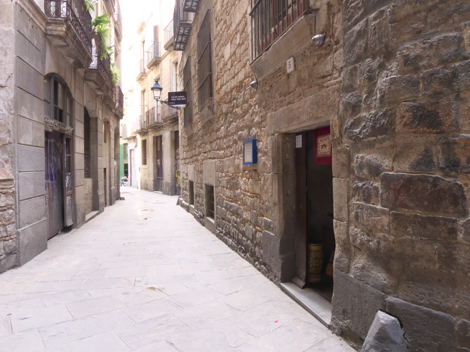 Calle estrecha y empedrada de casco antiguo, con fachadas de piedra y balcones de hierro, en la que se abre la puerta de acceso a un local con un tapiz rojo colgado en el quicio.