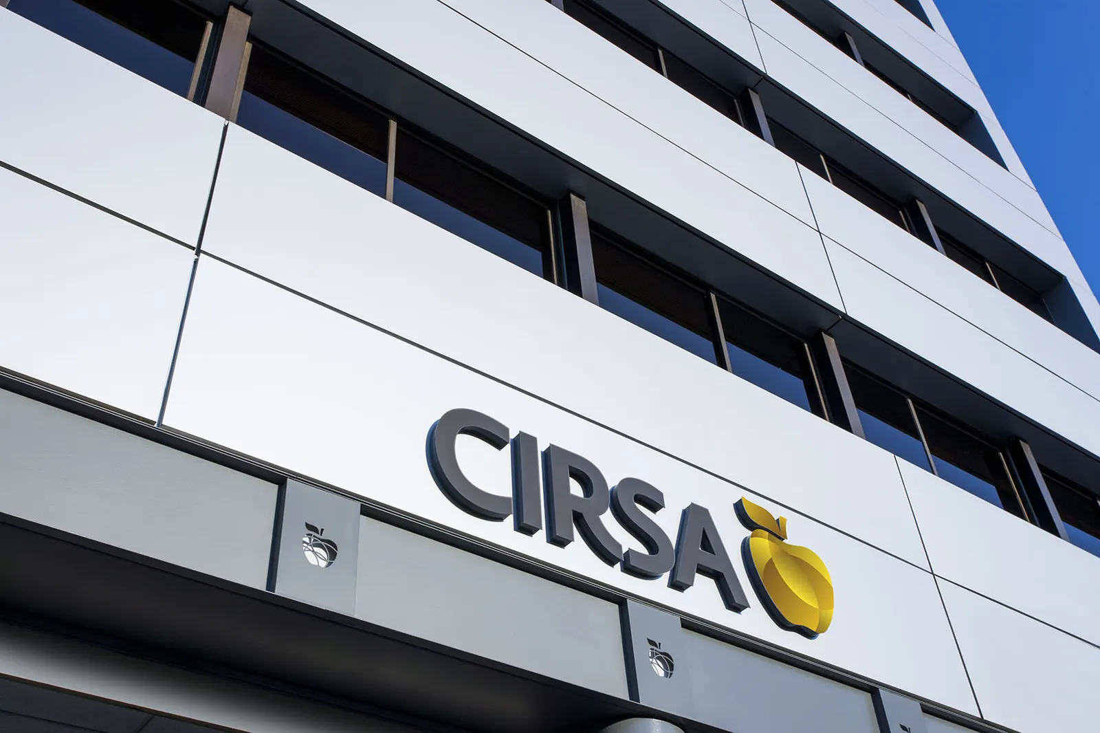 Fachada blanca de un edificio de oficinas con el rótulo corporativo de Cirsa y su logotipo, una manzana amarilla, bajo un cielo azul.