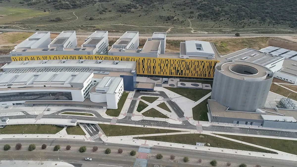 Vista aérea del Hospital Universitario de Toledo: un complejo blanco de varios bloques unidos por un cuerpo alargado de fachada amarilla, con un edificio circular gris a la derecha y carreteras en primer plano.
