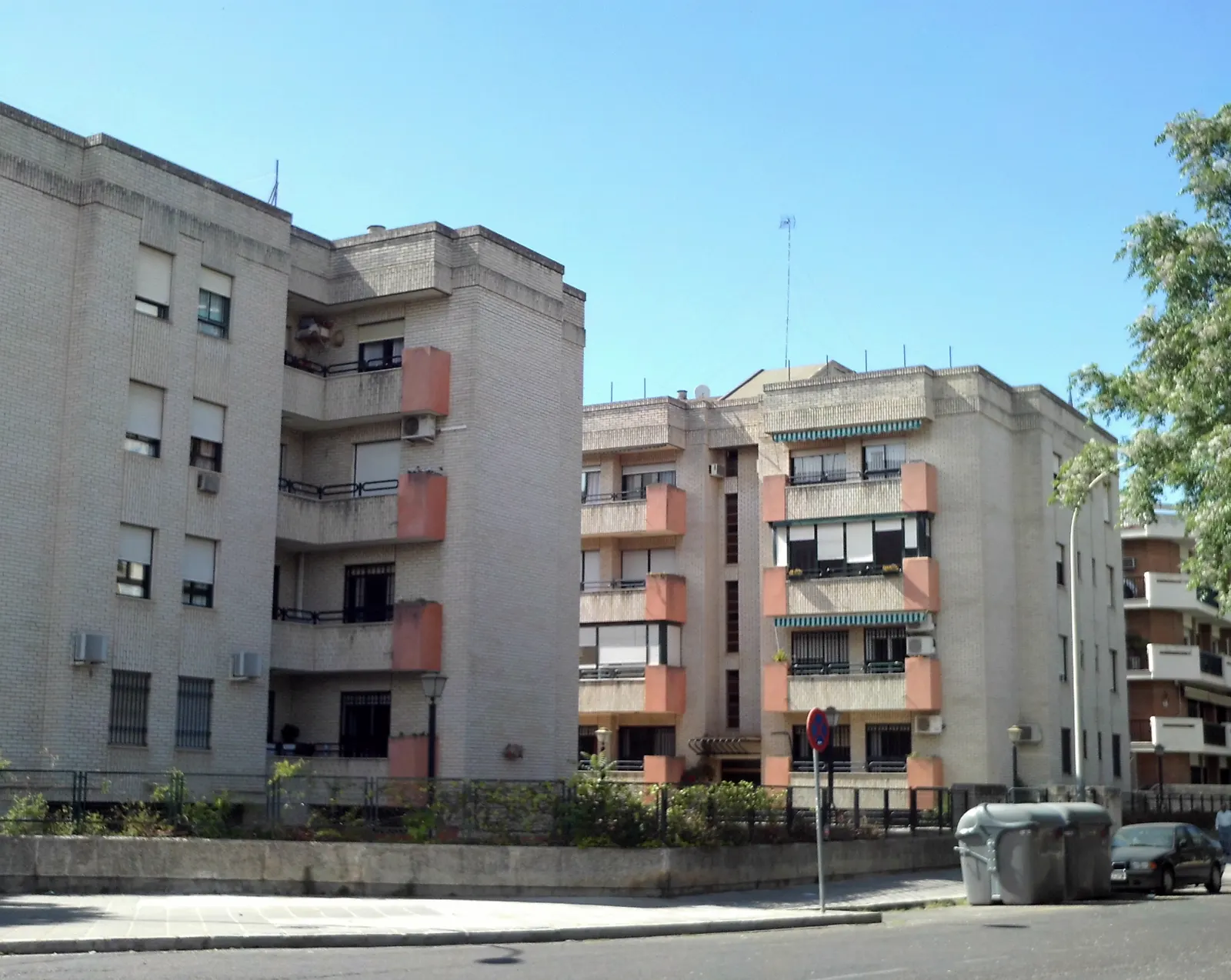 Dos bloques de pisos de cuatro alturas, de ladrillo claro con paños rojizos, con balcones, toldos y aparatos de aire acondicionado en las fachadas, junto a una calle vacía con un coche aparcado y contenedores de basura, bajo un cielo azul despejado.
