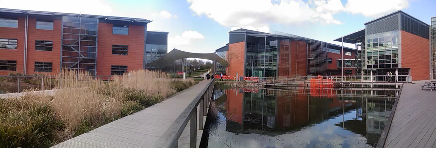 Edificios de oficinas de ladrillo rojo y grandes ventanales acristalados a ambos lados de un estanque, con pasarelas de madera y varias personas caminando, en el campus de la sede del grupo Vodafone en Newbury (Reino Unido)