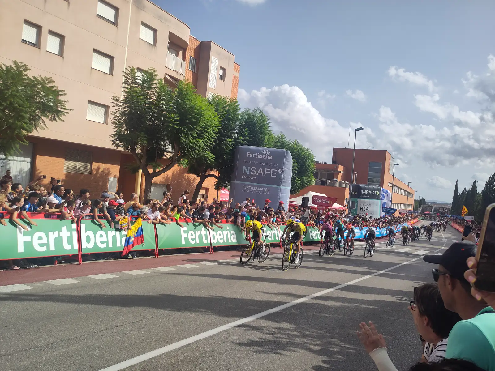 Dos ciclistas del Visma-Lease a Bike, con maillot amarillo y negro, encabezan el pelotón en una recta de meta flanqueada por vallas publicitarias verdes y público apretado contra ellas.