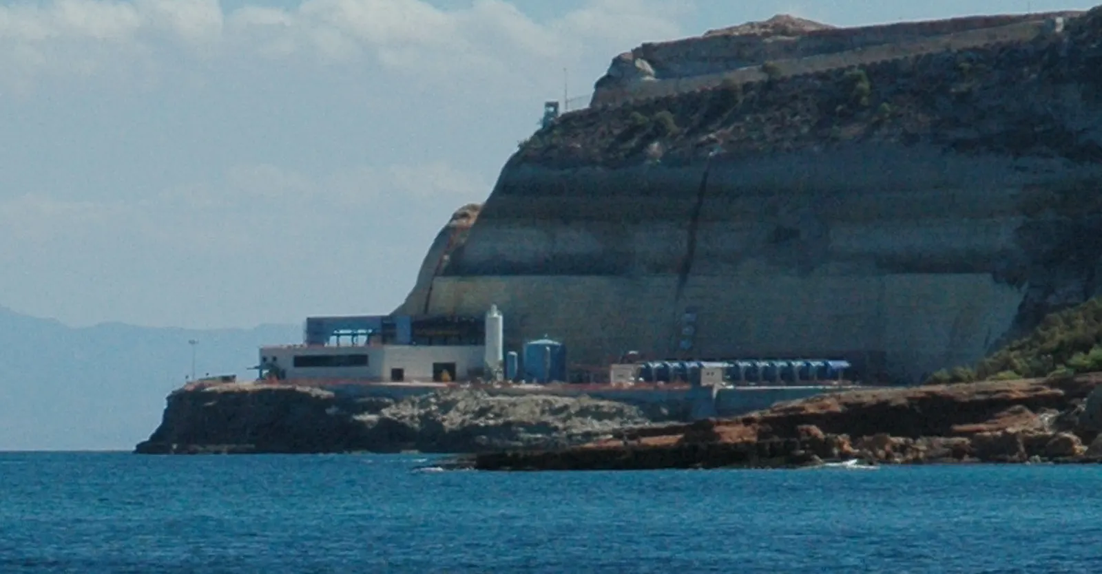 Planta industrial baja de color blanco y azul con un depósito cilíndrico y una hilera de equipos, encajada al pie de un acantilado de roca estratificada que cae al mar; agua azul en primer plano y costa brumosa al fondo.