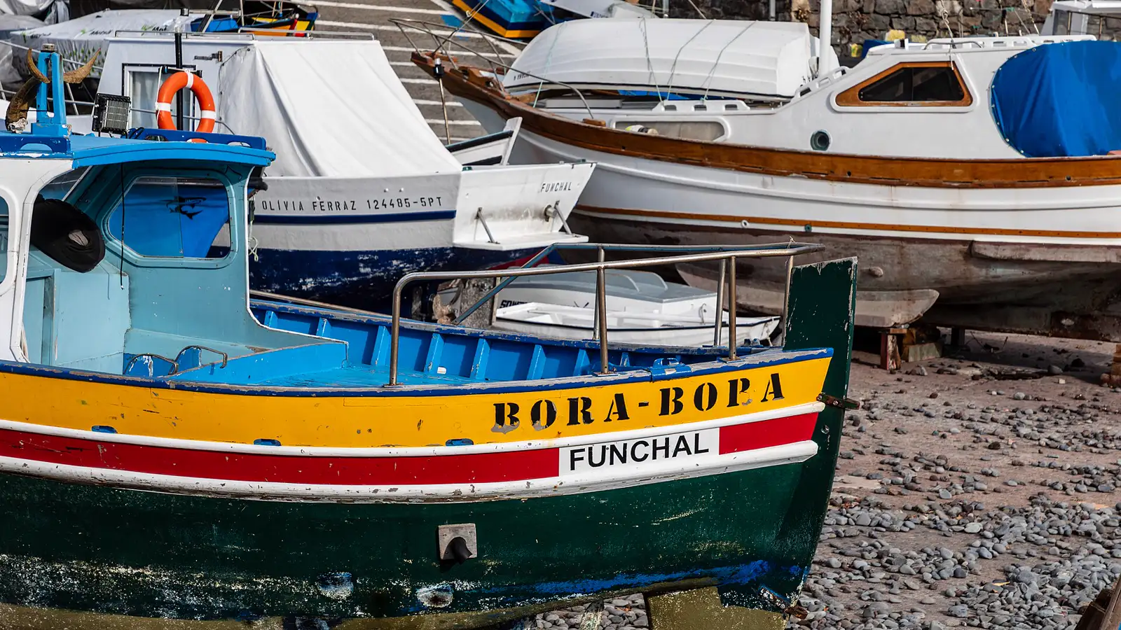 Barcas de pesca de madera pintadas de verde, amarillo, rojo y azul, varadas sobre la playa de cantos rodados del puerto de Câmara de Lobos, en Madeira; en la proa de la más cercana se lee «BORA-BOPA» y, debajo, el puerto de matrícula, «FUNCHAL».
