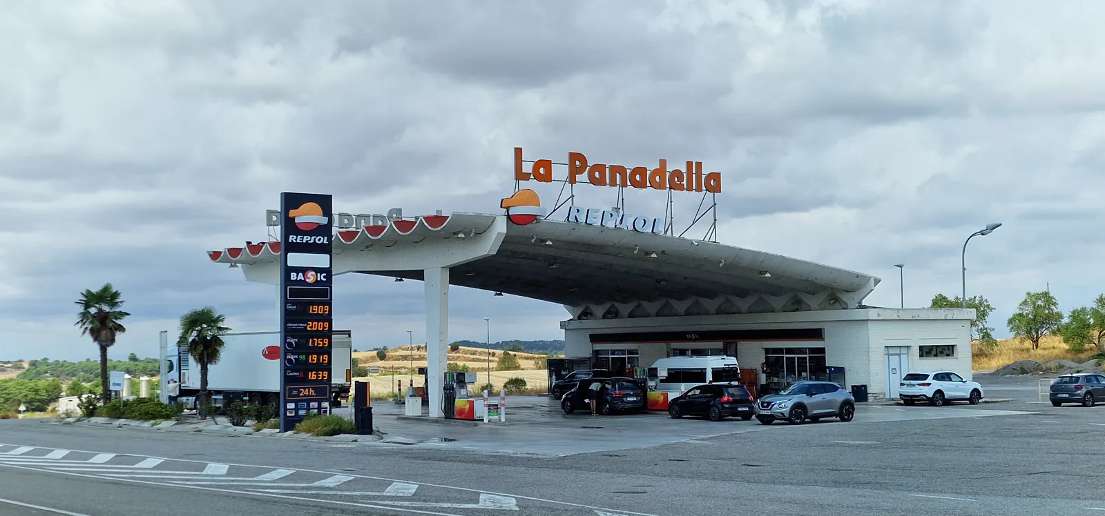 Estación de servicio Repsol junto a una carretera, con una gran marquesina de hormigón curvado y el rótulo «La Panadella». A la izquierda, un tótem de precios muestra el diésel a 1,909 euros, el diésel premium a 2,009, la gasolina 95 a 1,759, la 98 a 1,919 y el gasóleo B a 1,639. Bajo la marquesina reposta media docena de coches; al fondo, campos de rastrojo bajo un cielo nublado.