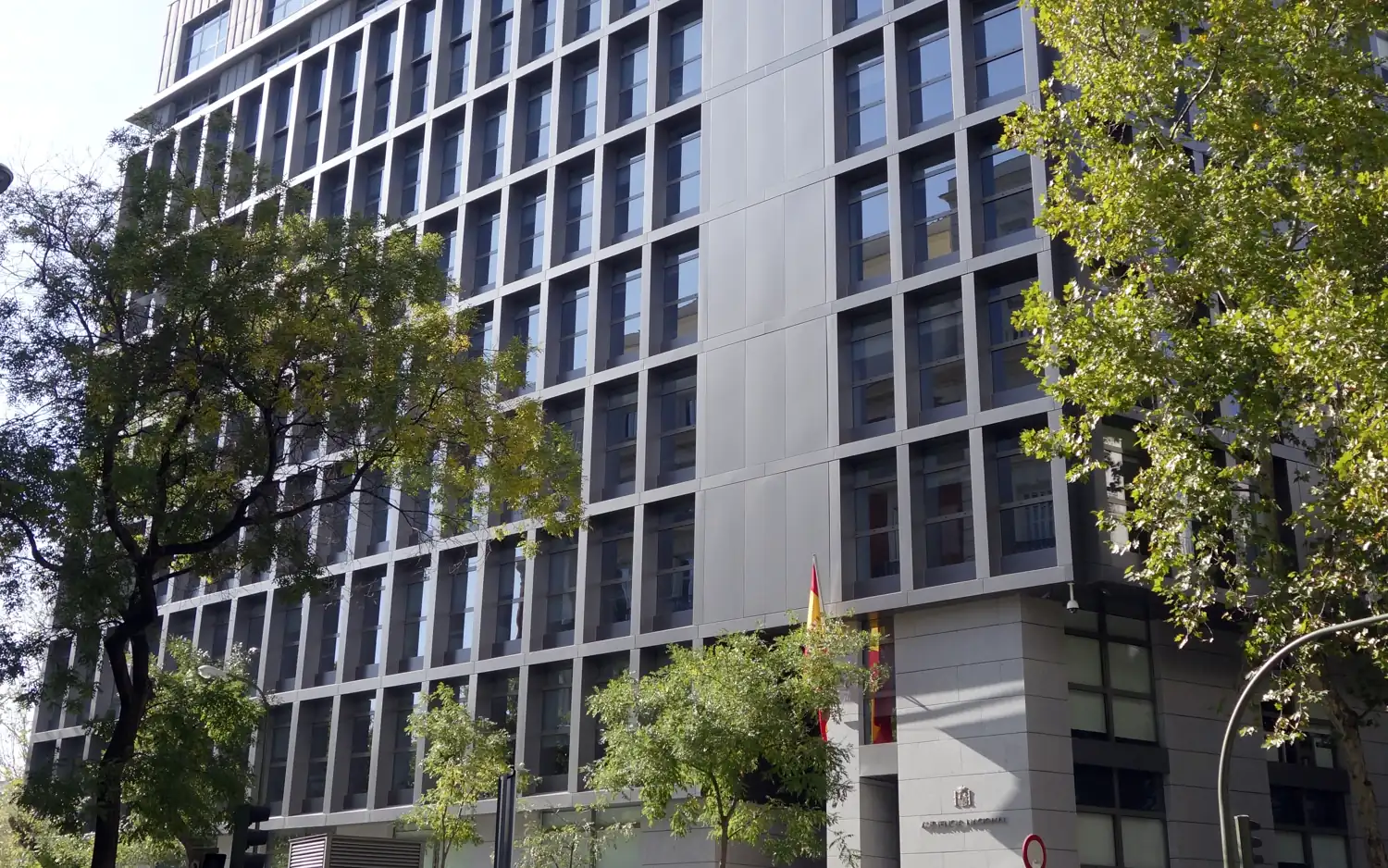 Fachada del edificio de la Audiencia Nacional en Madrid: un bloque moderno de varias plantas, con ventanas rectangulares enmarcadas en gris claro y árboles delante; junto a la entrada, la bandera de España y el escudo con el rótulo del tribunal.