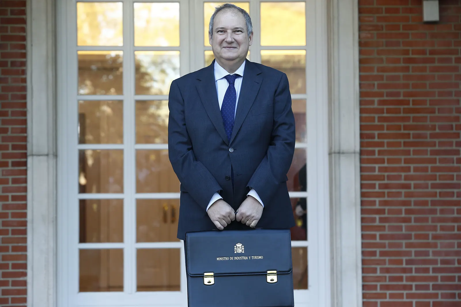 Hombre de traje azul marino y corbata azul de pie ante una puerta acristalada de marco blanco y muro de ladrillo, sosteniendo con las dos manos una cartera de cuero azul con el escudo de España y la leyenda «Ministro de Industria y Turismo».