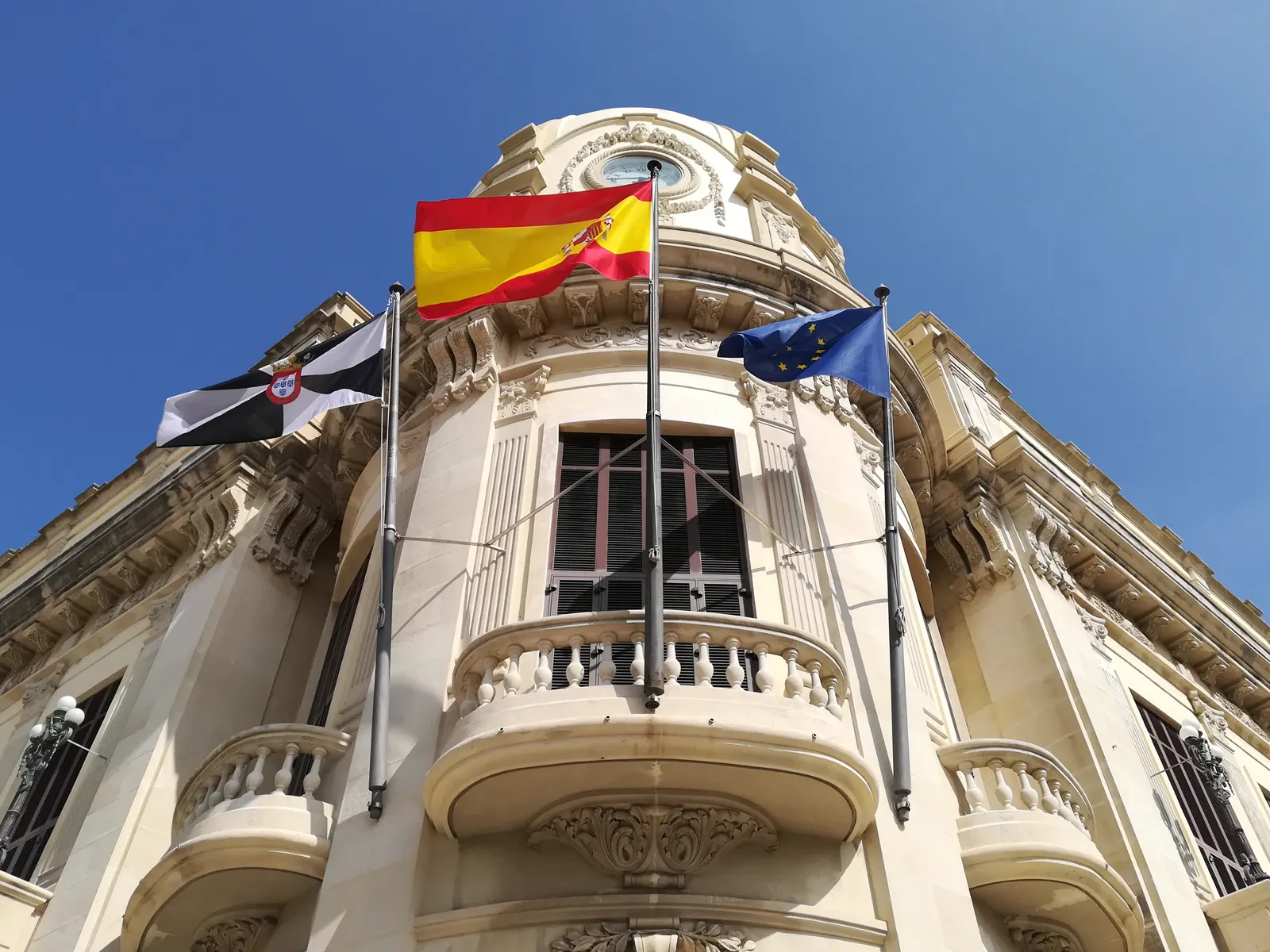 Esquina superior de un edificio institucional de piedra clara con balcón de balaustrada, fotografiada a contrapicado contra un cielo azul; ondean tres banderas: la de Ceuta, la de España y la de la Unión Europea.