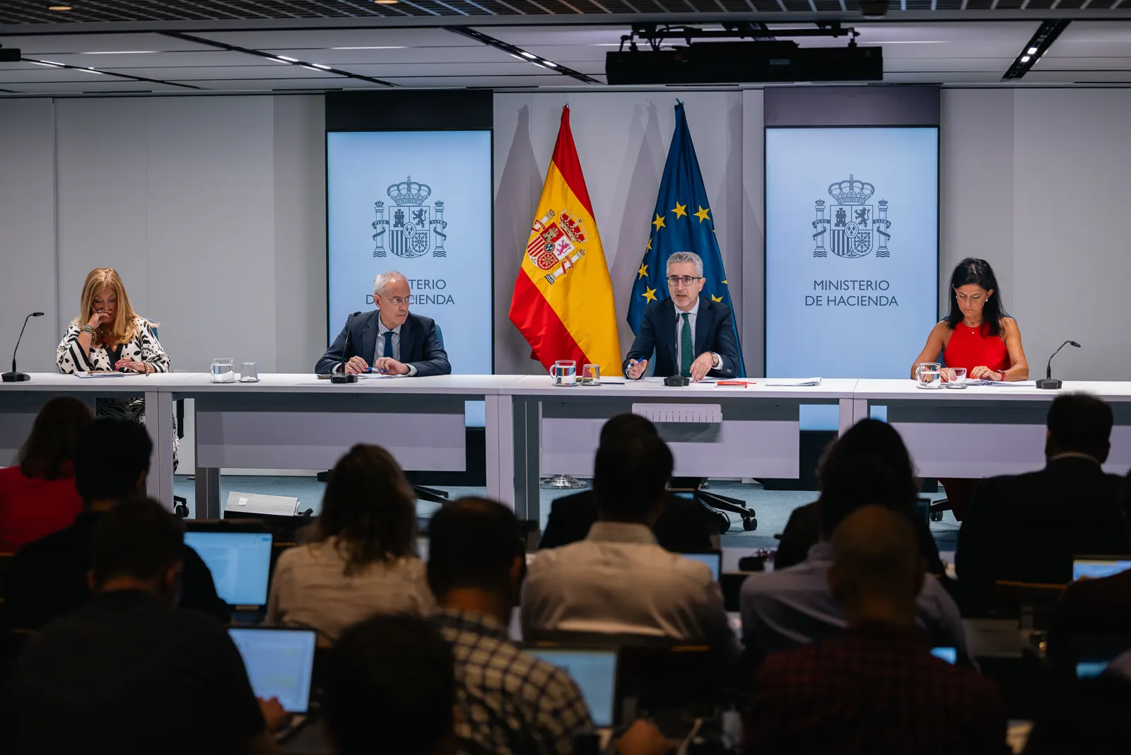 Cuatro personas sentadas tras una mesa alargada blanca, con micrófonos y documentos, ante paneles con el escudo de España y el rótulo «Ministerio de Hacienda»; entre ellas, las banderas de España y de la Unión Europea. En primer plano, de espaldas, periodistas con ordenadores portátiles.