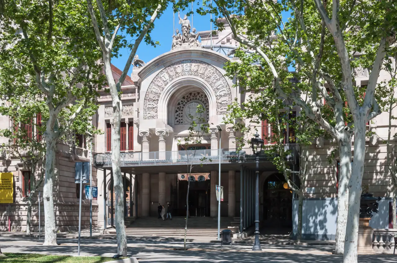 Fachada principal del Palacio de Justicia de Barcelona, edificio de piedra con un gran arco de medio punto sobre la entrada, vista entre plátanos del paseo de Lluís Companys.