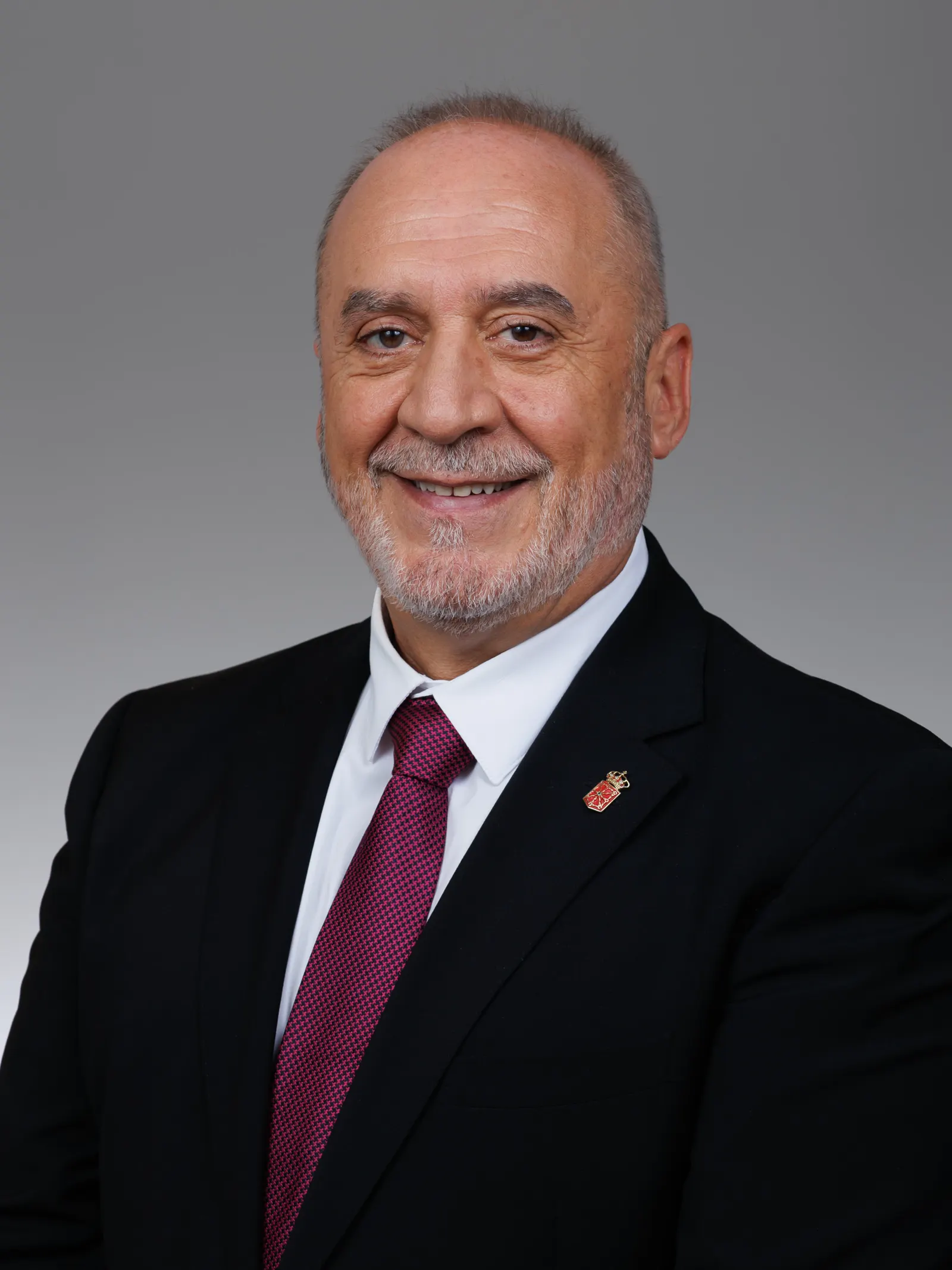 Retrato de estudio de un hombre con barba canosa y traje oscuro, camisa blanca y corbata granate, con un pin del escudo de Navarra en la solapa, sonriendo ante un fondo gris.