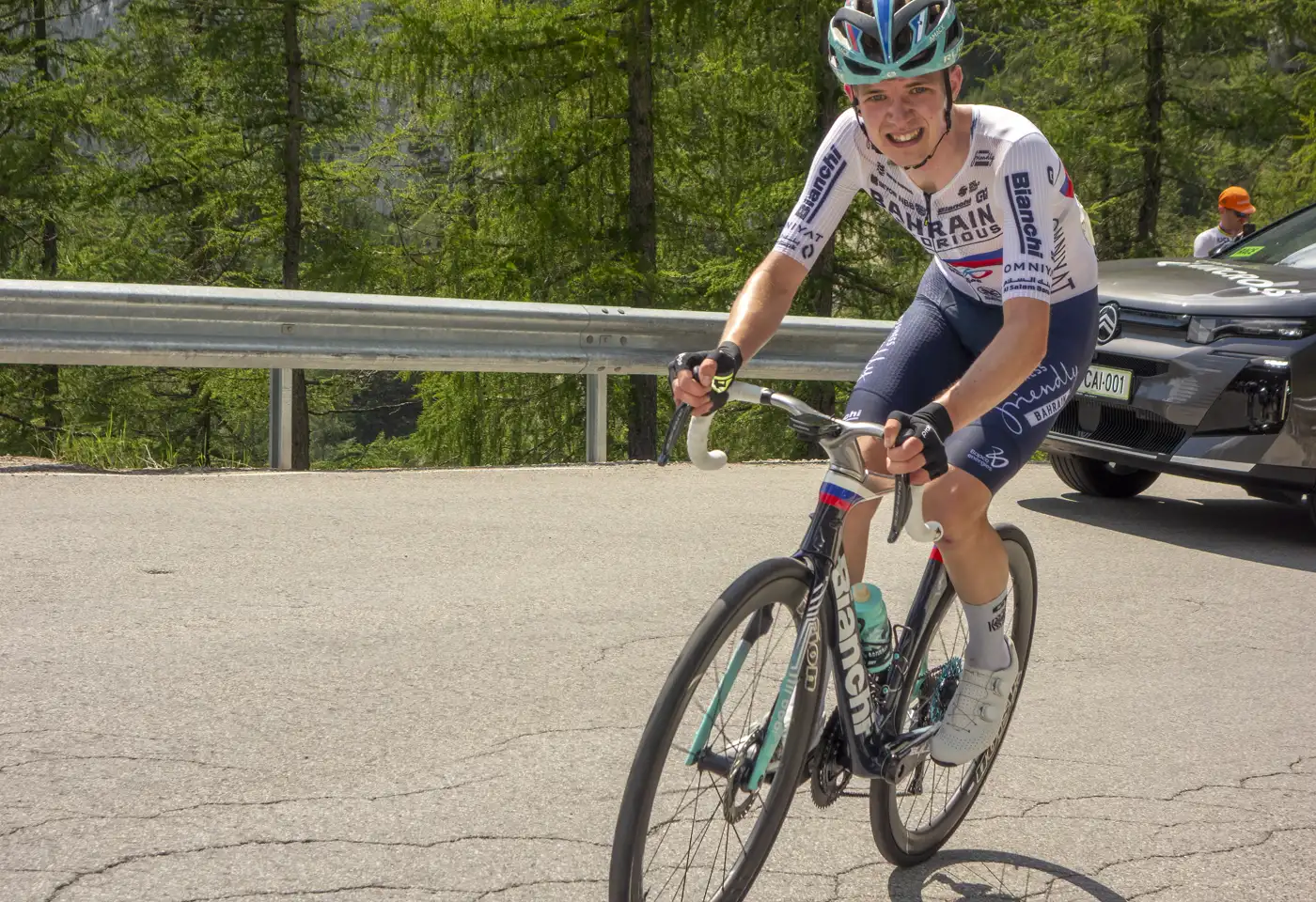 Un ciclista con maillot blanco y azul del Bahrain Victorious sube en pie sobre los pedales por una carretera de montaña, con bosque de coníferas y un coche de equipo detrás.