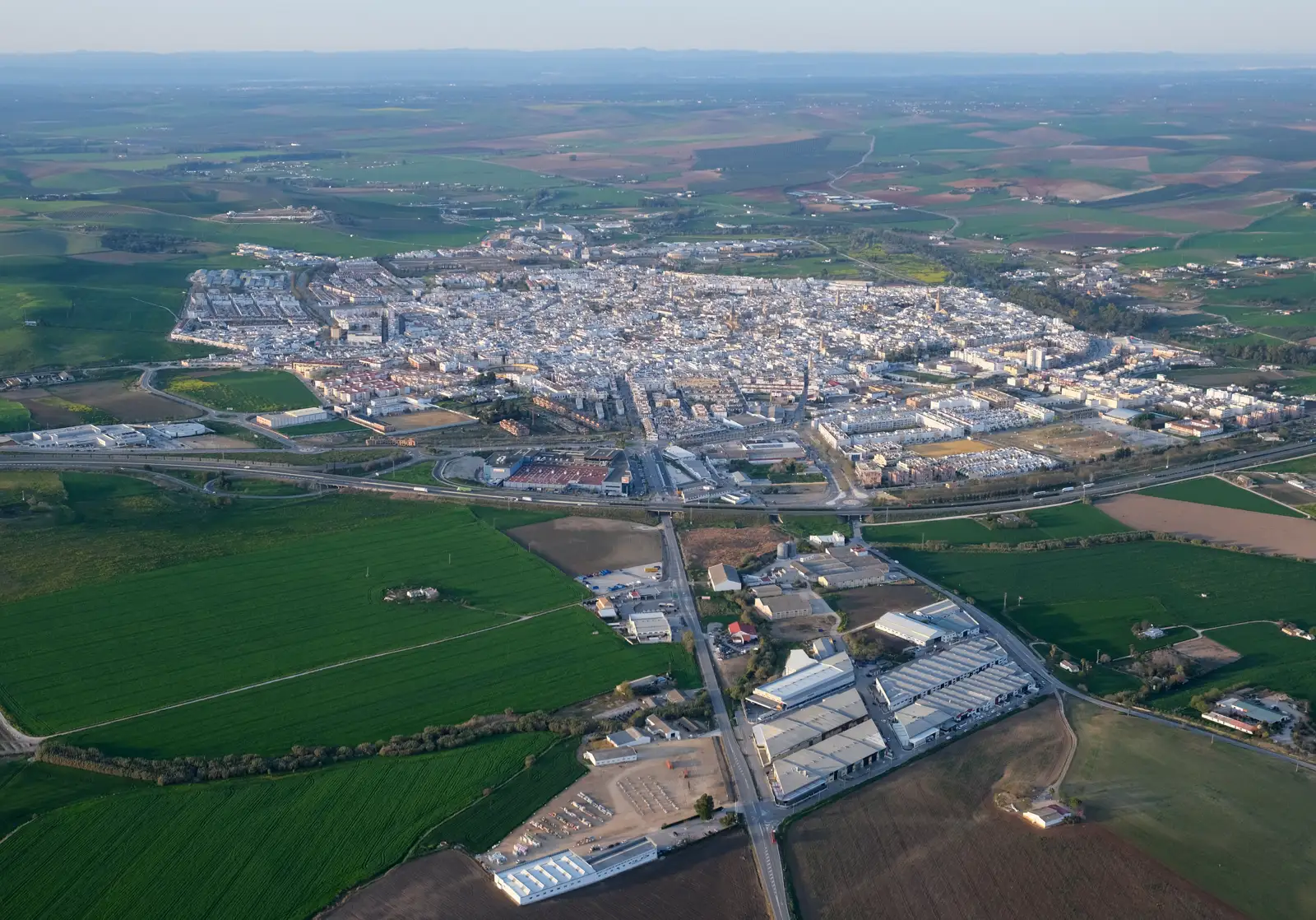 Vista aérea de Écija: el caserío blanco y compacto, con sus torres y su ronda de bloques, ocupa el centro de la imagen y a su alrededor se extiende hasta el horizonte una llanura de parcelas rectangulares de cultivo, verdes y pardas, cruzada por la autovía y por una carretera recta que baja hacia unas naves industriales en primer término.