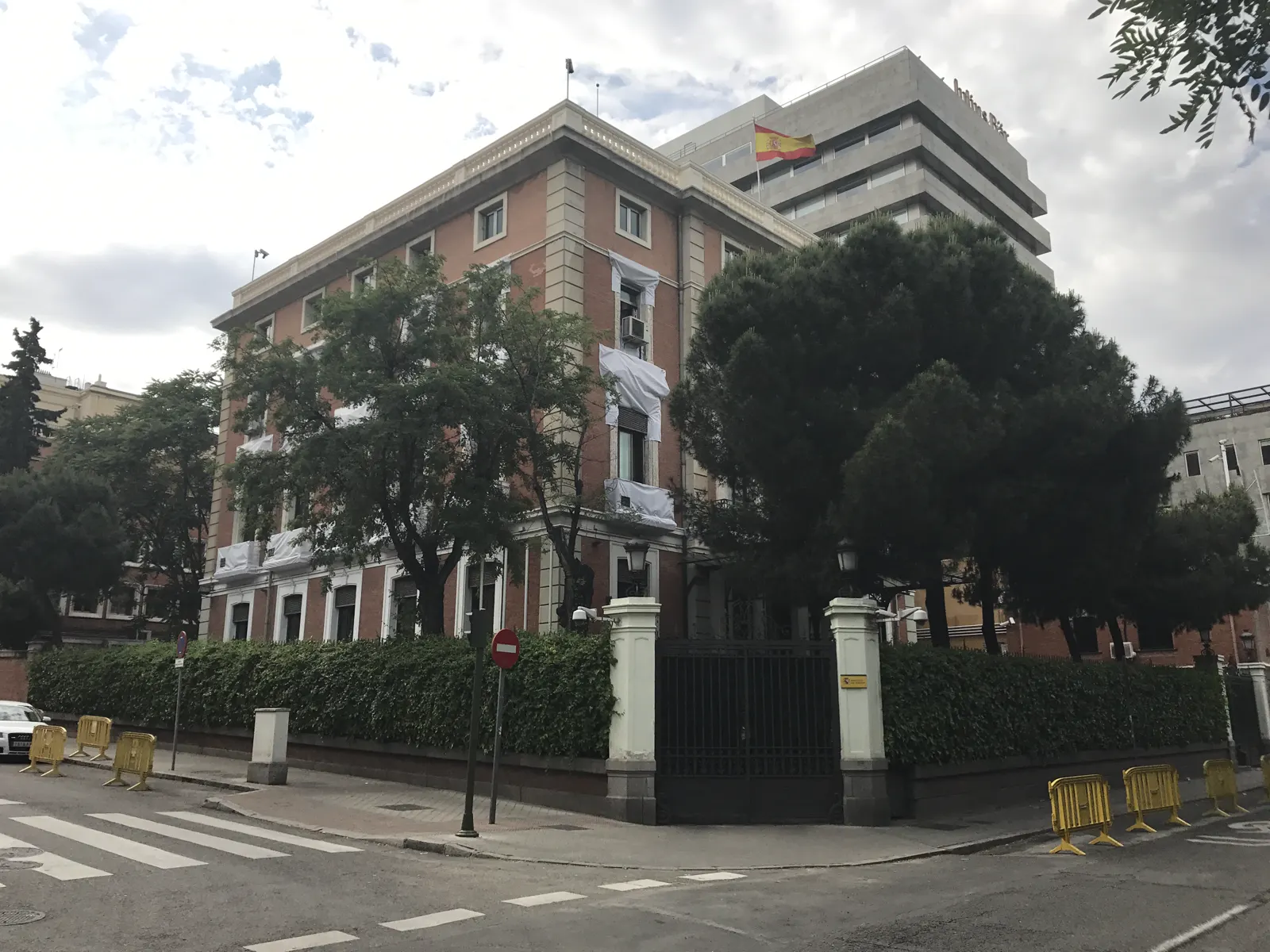 Esquina del palacio de los Condes de Casa Valencia, sede del Ministerio del Interior en Madrid: un edificio de cuatro plantas de ladrillo rojo con zócalo y cornisas de piedra clara, rodeado de árboles y de un seto tras una verja de hierro negro, con vallas amarillas en la acera y un paso de peatones en primer plano; al fondo ondea una bandera de España sobre un edificio contiguo.